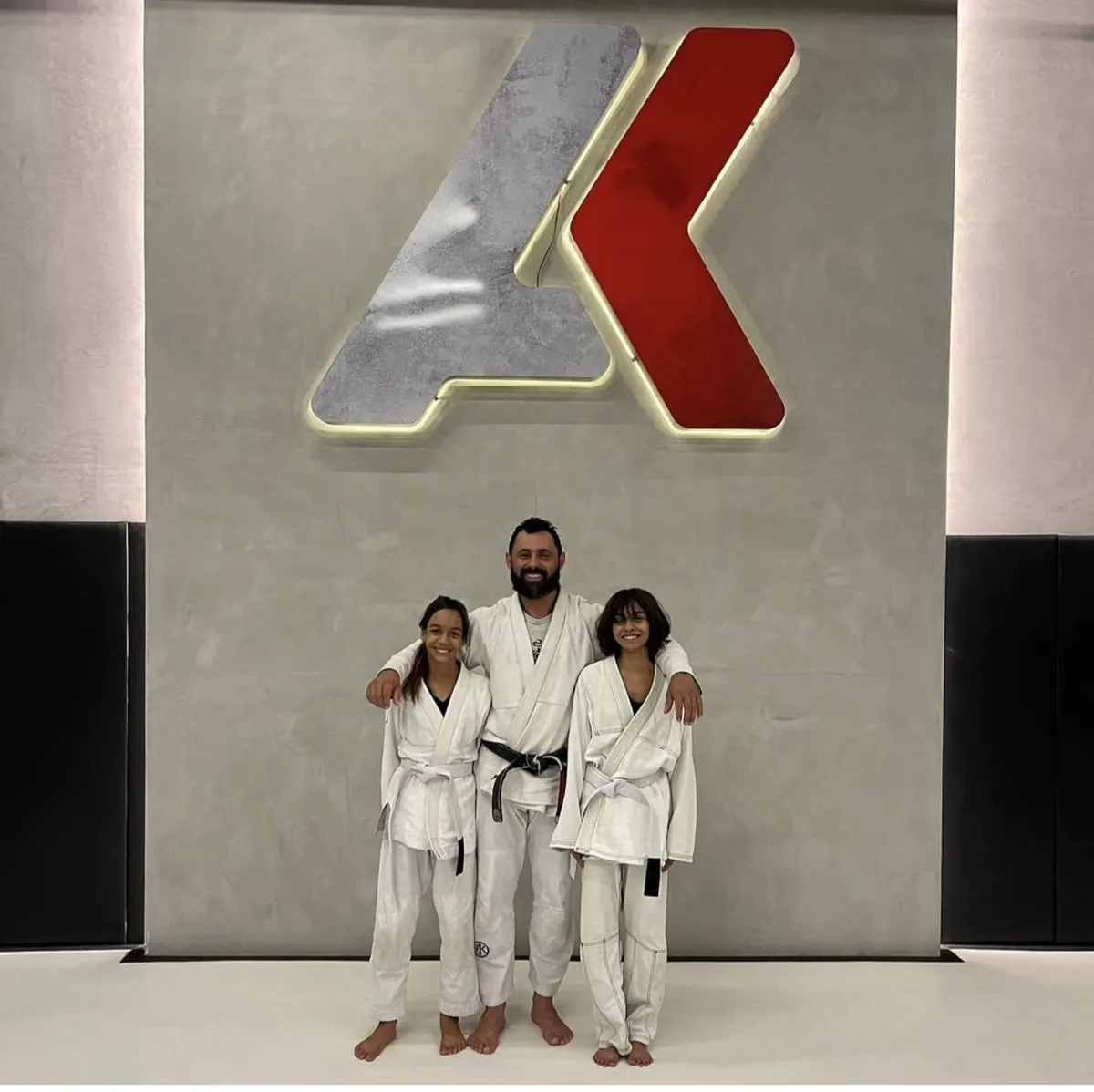 Profesor cinturón negro con dos alumnas en kimono frente al logotipo de la academia